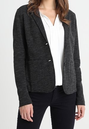 Nainen pukeutuneena teksturoituun tummanharmaaseen pukuun valkoisen paidan ja mustien housujen kanssa, seisoo kädet rentoina sivuillaan.