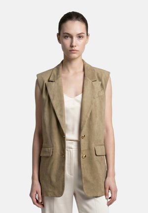 Donna che indossa un blazer senza maniche in camoscio color tan sopra una maglia di seta bianca e pantaloni, in piedi davanti a uno sfondo chiaro e semplice.