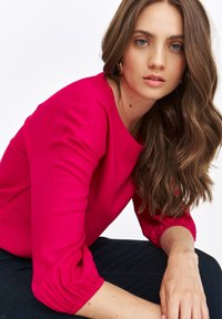 Fuksjowa bluzka z rękawami 3/4 o formie bufiastej, okrągłym dekoltem i teksturowanym materiałem, w zestawieniu z ciemnoniebieskimi dżinsami.