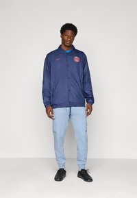 Navy blue, lightweight jacket with a collar and button closure; features red logo on the chest. Paired with light blue joggers and black shoes.