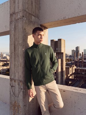 Jonge man in een groene jas met rits en beige broek leunt tegen een betonnen constructie op een dak met stadsgebouwen op de achtergrond.