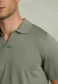Polo din bumbac tricotat verde, cu guler în V, mâneci scurte, țesătură texturată și cusături întărite, având un model delicat cu fâșii.