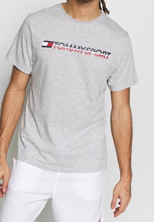 Mężczyzna w jasnoszarej koszulce Tommy Sport i białych szortach, stojący na jednolitym jasnym tle.