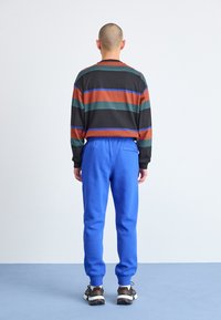 Striped long-sleeve shirt in black, green, red, and blue; blue sweatpants; black and white sneakers; viewed from behind against a plain background.