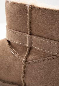 Detailní záběr na šití hnědé semišové boty a horní okraj s jemnou, světlou fleece podšívkou viditelnou.