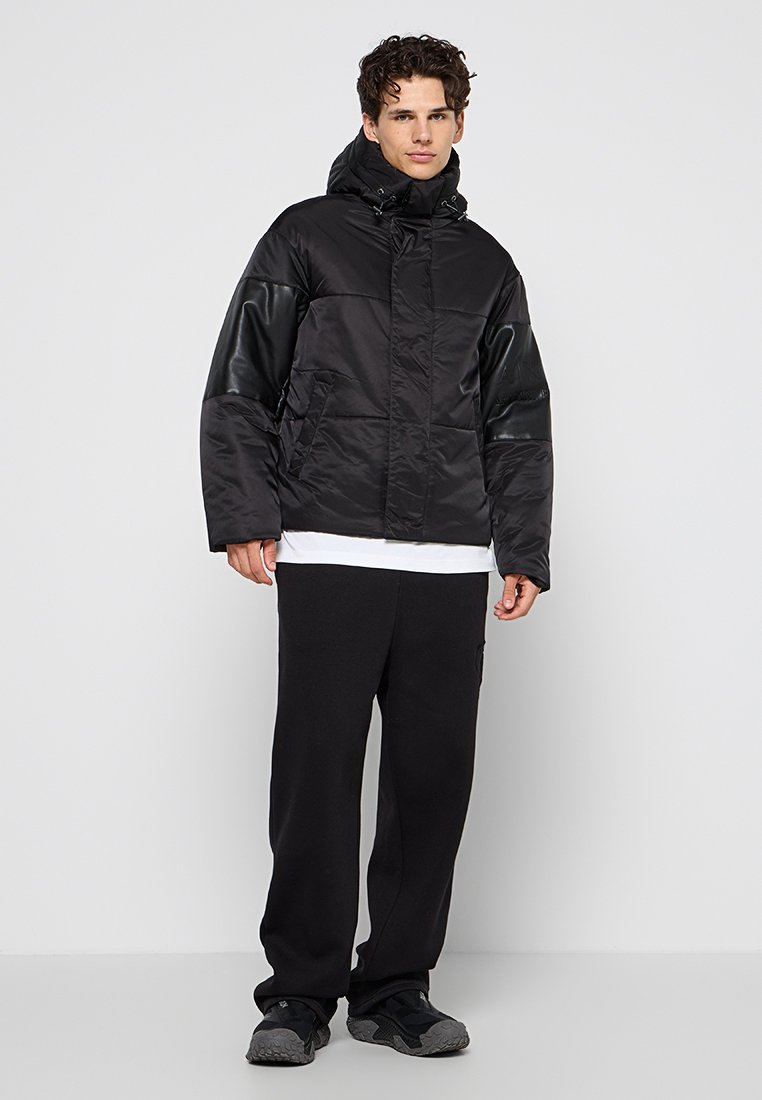 Giacca a vento nera con cappuccio, caratterizzata da una texture trapuntata, maniche in pelle a contrasto, tasche frontali e chiusura a bottoni a pressione. Abbinata a pantaloni sportivi neri.