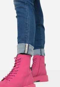 Růžové kotníkové boty na šněrování s hladkým povrchem, zesílenou špičkou a bočním zipem, doplněné o vyhrnuté tmavě modré džínové kalhoty.