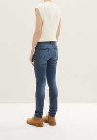 Bílý pletený bezramínkový top spárovaný s úzkými modrými džíny a béžovými kotníkovými botami. Pozadí má světlou neutrální barvu.