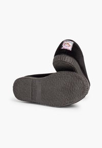 Paio di scarpe slip-on nere per bambini con suole testurizzate; una scarpa mostra l'etichetta con un cartone animato di una scimmia e il testo "Made in Spain".