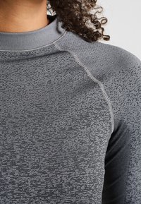 Szary, sportowy top z długim rękawem, wyróżniający się teksturowanym materiałem, rękawami raglanowymi oraz wysokim kołnierzem. Subtelna wariacja wzoru na całym materiale.