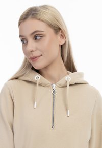 Felpa beige con zip frontale, dotata di cappuccio regolabile con cordoncino, tessuto testurizzato e dettagli in cerniera argentata. Design minimalista con tonalità neutre.