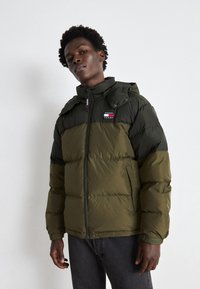 Chaqueta acolchada en verde y verde oscuro con cierre de cremallera, cuello alzado y parche de logo en el pecho; cuenta con bolsillos y capucha.