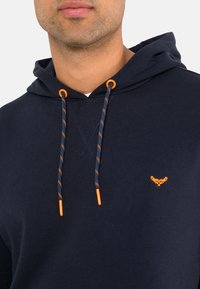 Felpa con cappuccio blu navy realizzata in tessuto morbido, con lacci arancioni a spirale e un piccolo logo ricamato in arancione sul petto.