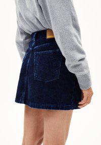 Marineblauwe corduroy mini-jurk met verticale ribbels, voorzakken en een bruine leren label op de tailleband; gecombineerd met een grijze trui.