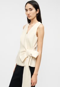 Κρεμ τοπ με λαιμόκοψη σε σχήμα V, κατασκευασμένο από απαλό ύφασμα, με λεπτομέρεια φιόγκου στη μέση. Το σχέδιο διαθέτει φαρδιές τιράντες και εφαρμοστό σχήμα.