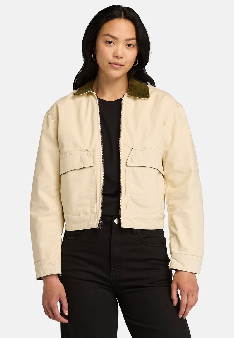 Klippt beige jacka i bomull med en grön kordfloretkrage, med två framfickor och dragkedjestängning. Bärs över en svart topp.