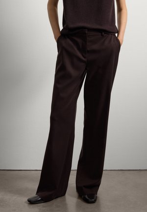 Persona con pantalones anchos negros y zapatos negros, de pie con las manos en los bolsillos contra una pared clara y lisa.