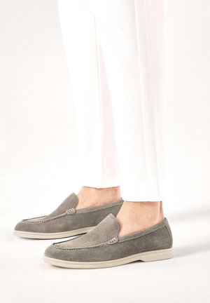 Person trägt hellgraue Wildleder-Loafer mit weißen Sohlen und weißer Hose, steht vor neutralem Hintergrund.