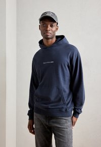 Hanorac cu glugă de culoare bleu marin, realizat dintr-un material moale, având un buzunar tip kengur și text logo în față. Este asortat cu pantaloni din denim gri și o șapcă.