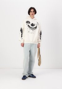 Hvit hettegenser med svart grafisk design, frontlomme, kombinert med lyseblå jeans og marineblå sneakers. Holder en lys brun skulderveske.
