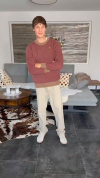 Red patterned sweater with navy accents, beige trousers, and white sneakers. Indoor setting with a dark tile floor and modern furniture.