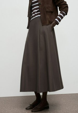 Persona con chaqueta marrón de ante, camisa a rayas, falda larga acampanada gris y bailarinas marrón oscuro, parada sobre un suelo salpicado.