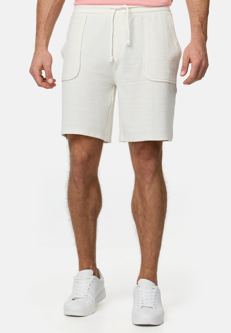 Pantalones cortos blancos texturizados con dos bolsillos frontales y cintura ajustable con cordón, combinados con zapatillas blancas.