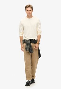 Roomwitte lange mouwen shirt, tan corduroy broek en een blauw-groen geruit shirt om de taille gebonden. Zwarte schoenen met een glanzende afwerking.