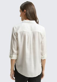 Blouse de couleur claire avec des manches retroussables, tissu texturé, empiècement au dos et motif rayé subtil. Design d'ourlet incurvé pour un ajustement décontracté.