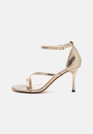 Sandale à talons hauts dorée avec une fine bride à la cheville, bout ouvert et motif peau de serpent texturé sur fond blanc.