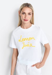 White cotton t-shirt featuring yellow handwritten text "Lemon Juice." Short sleeves and a crew neckline with a relaxed fit.