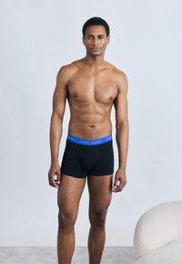 Μαύρα boxer εσώρουχα με μπλε ελαστική μέση που φέρει το λογότυπο. Ο τόνος του δέρματος είναι ορατός, ενώ το μοντέλο στέκεται μπροστά σε ανοιχτό γκρι φόντο.