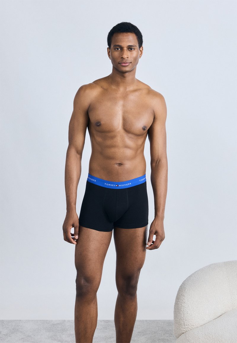 Μαύρα boxer εσώρουχα με μπλε ελαστική μέση που φέρει το λογότυπο. Ο τόνος του δέρματος είναι ορατός, ενώ το μοντέλο στέκεται μπροστά σε ανοιχτό γκρι φόντο.