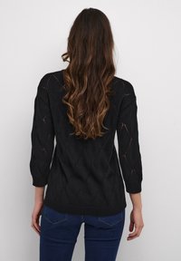 Schwarzer Strickpullover mit Rautenmuster, Dreiviertelärmeln und Ribbenbund. Getragen mit dunklen Jeans, strukturierter Stoff sichtbar.