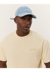 Tricou galben deschis cu un logo brodat; asortat cu o șapcă de baseball din suede albastru, având o margine curbată și o curea ajustabilă.