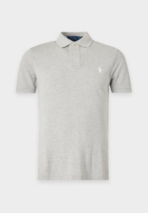 Homme portant des lunettes de soleil noires, un polo gris clair, un short beige longueur genou, des chaussettes blanches et des baskets beiges, debout avec une main dans la poche.