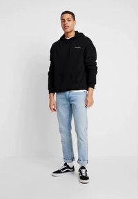 Schwarzer Hoodie mit Fronttasche und Zugschnur, über einem weißen Hemd getragen. Hellblaue Jeans und schwarze Sneakers vervollständigen das Outfit.