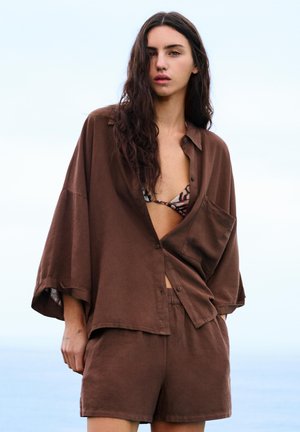 Jeune femme aux longs cheveux foncés portant une chemise marron ample, un short assorti et un haut de bikini à motifs, debout à l'extérieur au bord de la mer.