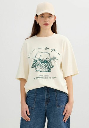 Mujer joven con una camiseta oversize color crema con gráfico de cesta de picnic y texto, vaqueros azules, gorra beige y pendientes de aro dorados.