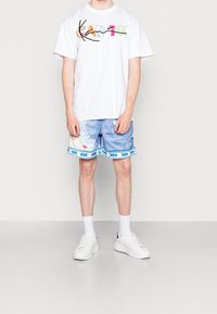 Camiseta blanca con gráfico multicolor "KAN", combinada con pantalones cortos de baño azules con estampado y un ribete decorativo, junto con zapatillas blancas.