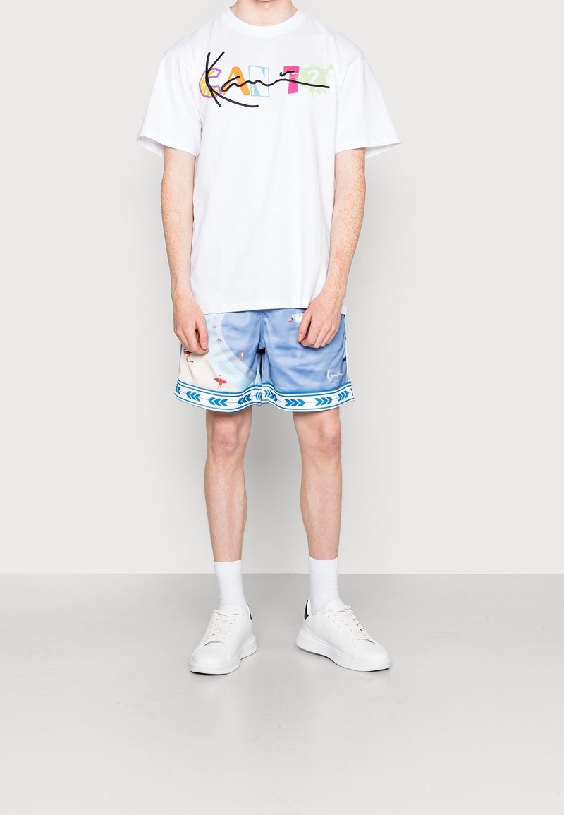 Camiseta blanca con gráfico multicolor "KAN", combinada con pantalones cortos de baño azules con estampado y un ribete decorativo, junto con zapatillas blancas.