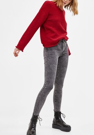 Femme portant un pull rouge, un jean skinny gris délavé et des bottes à plateforme noires à lacets, posant devant un fond blanc uni.