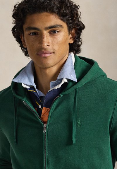 Camisola verde com fecho de correr e cordões, tecido texturizado, logótipo no lado esquerdo do peito. Usada por cima de uma camisa azul e branca com gola e detalhes coloridos.