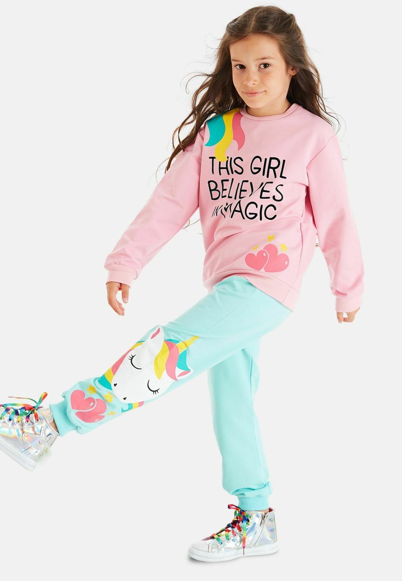 Rosa sweatshirt med svart text, tillsammans med mintgröna joggers som visar en enhörningsgrafik. Texten lyder "DENNA TJEJ TROR PÅ MAGI." Silverfärgade sneakers.