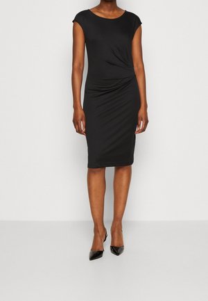 Frau trägt ein figurbetontes, knieumspielendes schwarzes Kleid mit kurzen Ärmeln und seitlichen Raffungen, kombiniert mit schwarzen spitzen Absatzschuhen.