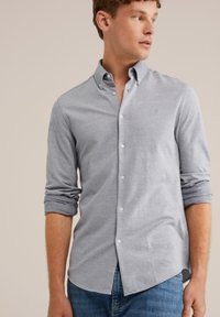 Joven vestido con una camisa de botón gris claro con mangas enrolladas y jeans azules, mirando hacia su derecha contra un fondo neutro.