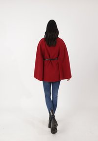 Mantello rosso con cintura in vita, realizzato in materiale spesso. Abbinato a jeans aderenti blu e stivaletti neri alla caviglia. Tessuto liscio e design strutturato.