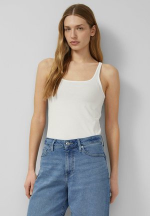 Młoda kobieta z długimi jasnobrązowymi włosami, ubrana w biały top i niebieskie dżinsy z wysokim stanem, stoi na tle jednolitego szarego tła.