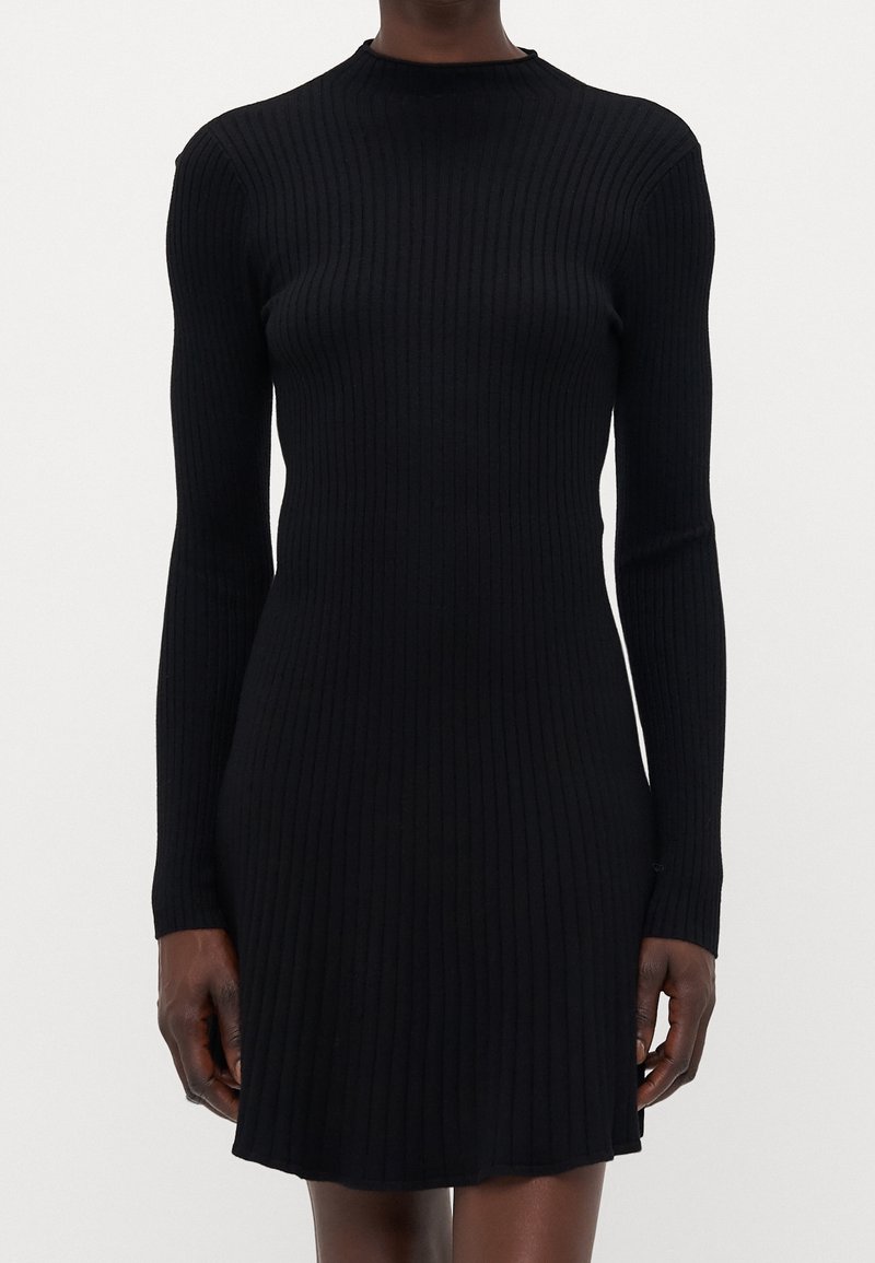 Femme vêtue d'une robe noire moulante en maille côtelée à manches longues avec une jupe courte, debout devant un fond clair uni.