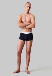 Boxeri scurți din bumbac de culoare bleumarin cu un tiv alb cu logo-ul Calvin Klein. Modelul stă cu brațele încrucișate, evidențiind croiala și designul.